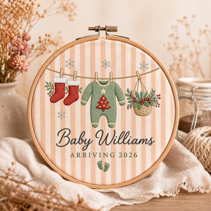 May include: A round wooden embroidery hoop featuring a light peach and white striped fabric background. The design includes embroidered red stockings, a green baby onesie with a red Christmas tree, and a basket of holly. Text reads Baby Williams ARRIVING 2026.
