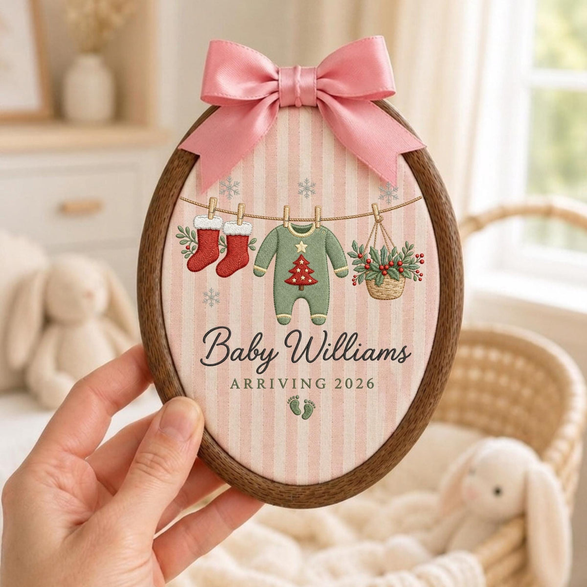 May include: An oval wooden embroidery hoop containing fabric with pink vertical stripes. It features a pink satin bow at the top and an illustration of a clothesline with two red stockings, a green baby onesie with a tree, and a basket of holly.
