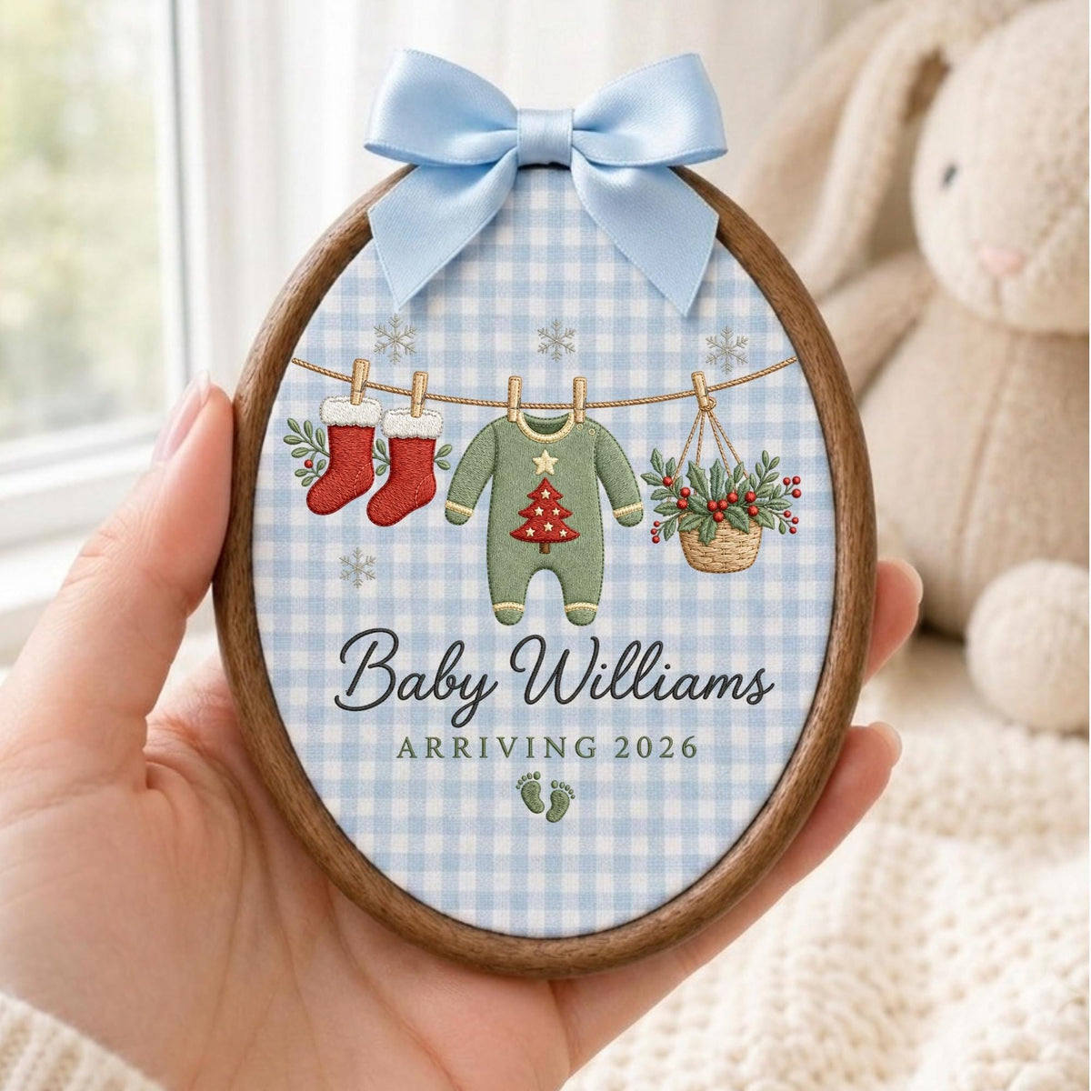 May include: An oval wooden embroidery hoop frame featuring a light blue gingham fabric background. The design includes a clothesline with two red stockings, a green baby onesie with a red Christmas tree, and a basket of holly. Text reads Baby Williams ARRIVING 2026 with small green footprints and a light blue satin bow.