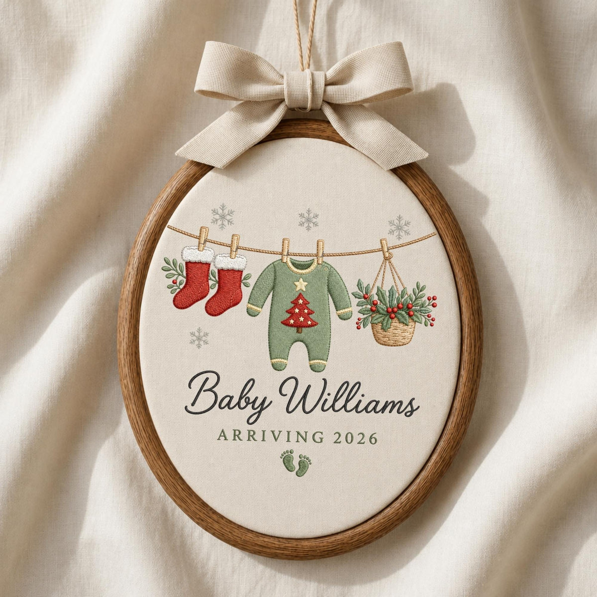 May include: An oval wooden embroidery hoop ornament featuring a beige fabric center with a beige ribbon bow at the top. The design shows two red stockings, a green baby onesie with a red tree, and a basket of holly hanging on a line with snowflakes.