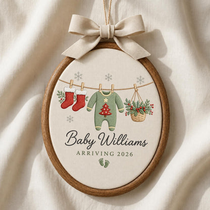 May include: An oval wooden embroidery hoop ornament featuring a beige fabric center with a beige ribbon bow at the top. The design shows two red stockings, a green baby onesie with a red tree, and a basket of holly hanging on a line with snowflakes.