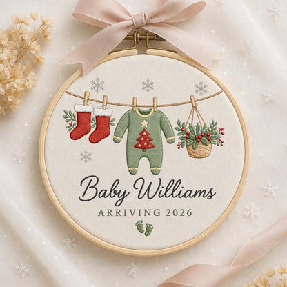 May include: A round wooden embroidery hoop featuring a cream fabric background with a light pink ribbon bow at the top. The design shows a clothesline with two red stockings, a green baby onesie with a red Christmas tree, and a basket of holly. Text reads Baby Williams ARRIVING 2026 with green baby footprints.