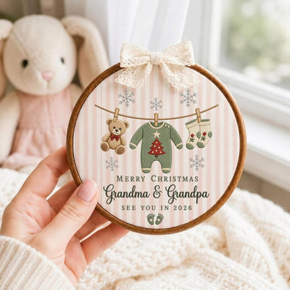 May include: A round wooden embroidery hoop frame containing fabric with a vertical pink and white striped pattern. It features a cream lace bow at the top and a clothesline graphic with a teddy bear, a green onesie, and socks. Text reads Merry Christmas Grandma &amp; Grandpa See You In 2026.