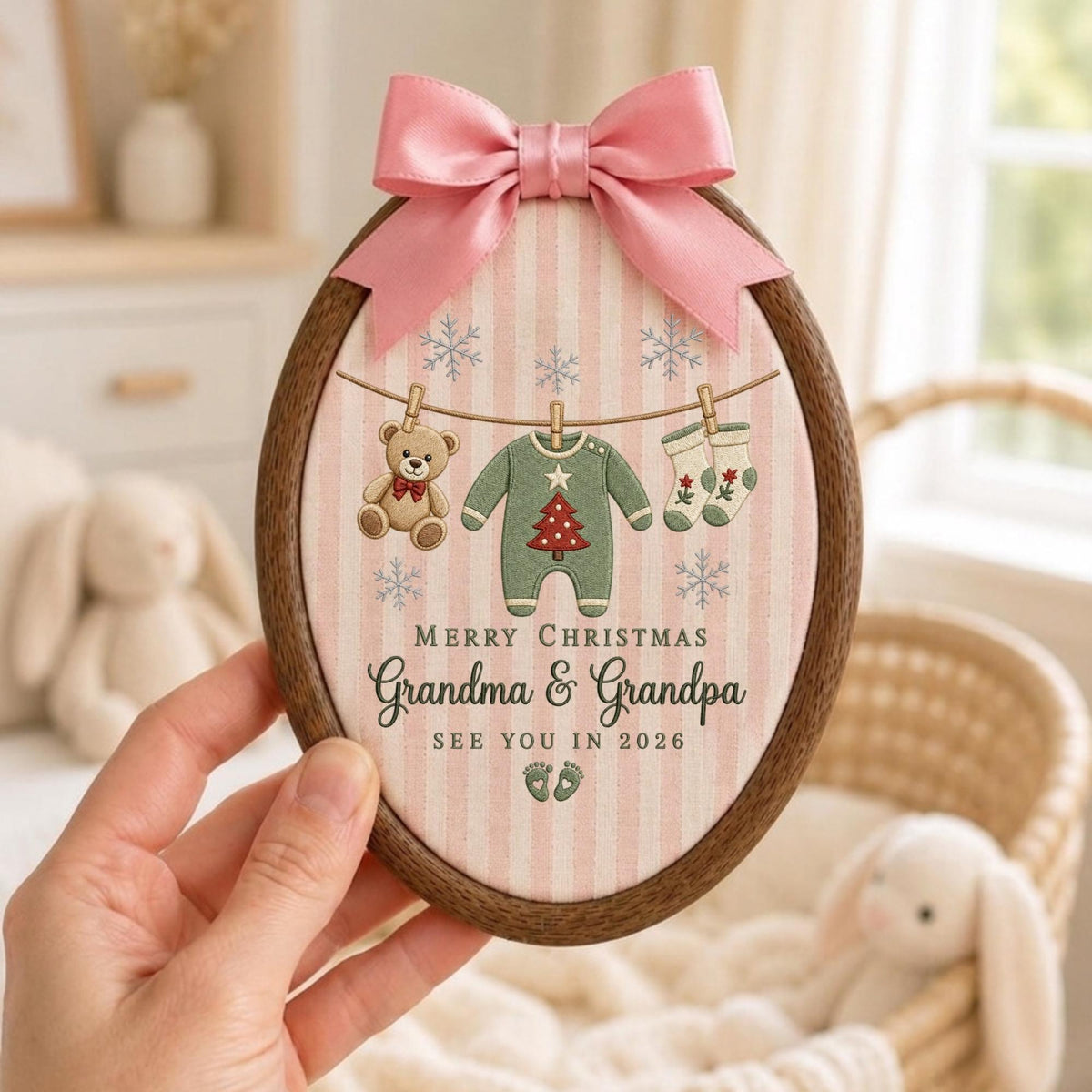 May include: An oval wooden embroidery hoop frame containing fabric with a pink and white striped pattern. The fabric features a clothesline with a teddy bear, a green onesie with a red Christmas tree, and socks. Text reads: MERRY CHRISTMAS Grandma &amp; Grandpa SEE YOU IN 2026, with a pink satin bow on top.