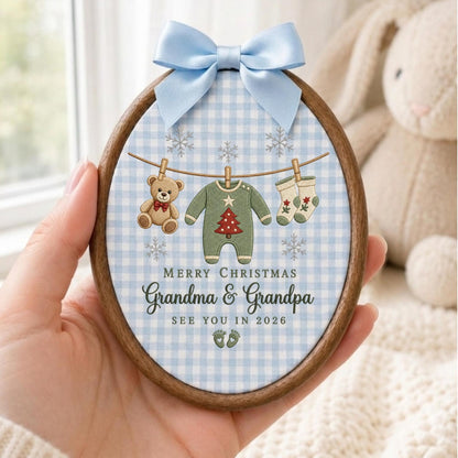 May include: An oval wooden embroidery hoop ornament featuring a light blue and white gingham fabric background. A light blue satin bow sits at the top. The fabric displays a clothesline with a teddy bear, a green onesie, and socks, with the text MERRY CHRISTMAS Grandma &amp; Grandpa SEE YOU IN 2026 and two small footprints.