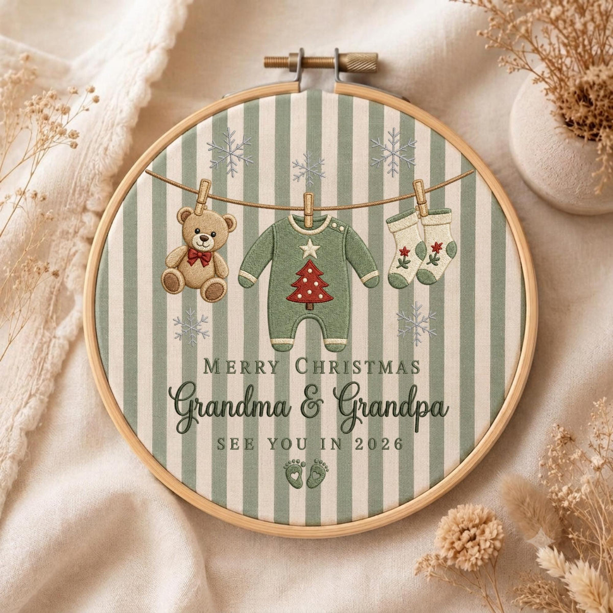May include: A round wooden embroidery hoop containing fabric with green and white vertical stripes. The design features a teddy bear, a baby onesie with a red Christmas tree, and two socks hanging on a line, with the text Merry Christmas Grandma &amp; Grandpa See You In 2026 and two small footprints.