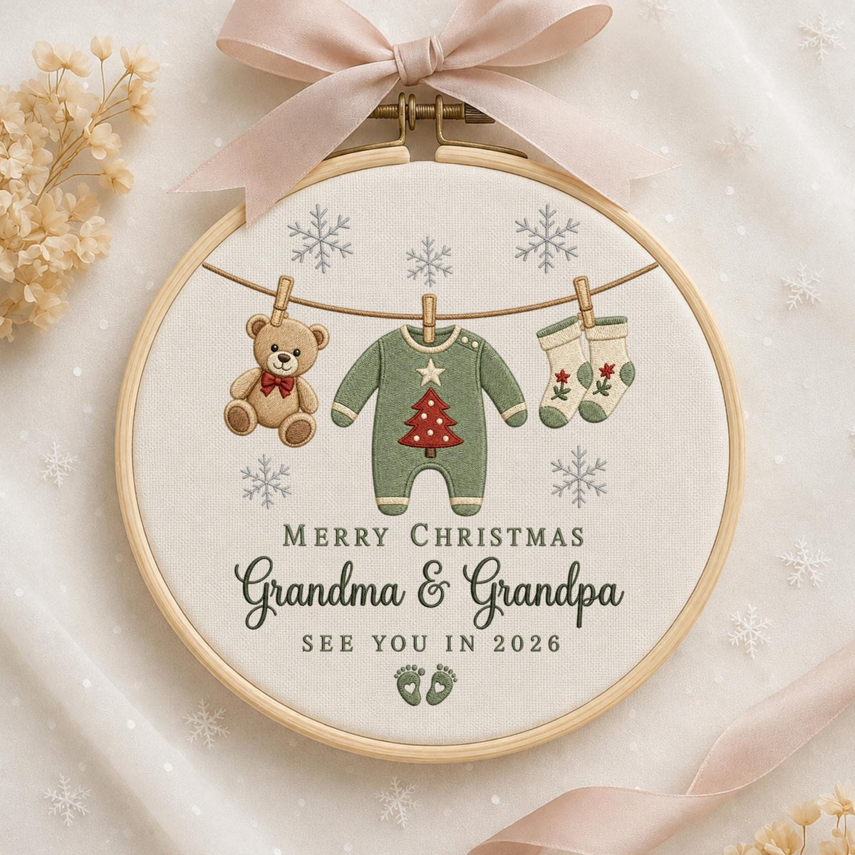 May include: A round wooden embroidery hoop containing fabric with a light beige background. The design features a clothesline with a teddy bear, a green baby onesie with a red Christmas tree, and two socks. Text reads: MERRY CHRISTMAS Grandma &amp; Grandpa SEE YOU IN 2026, with baby footprints and snowflakes.