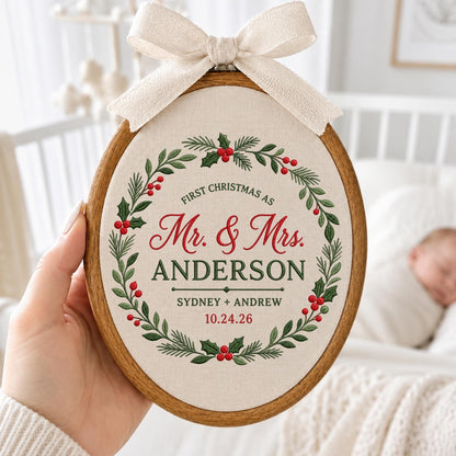 May include: An oval wooden embroidery hoop containing cream fabric with a printed Christmas wreath design. The text reads: FIRST CHRISTMAS AS Mr. &amp; Mrs. ANDERSON SYDNEY + ANDREW 10.24.26. A beige fabric bow is attached to the top of the hoop.