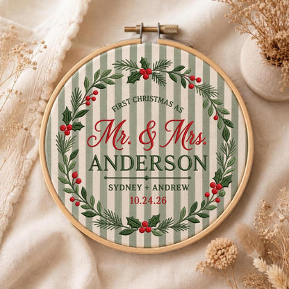 May include: A circular embroidery hoop containing fabric with vertical sage green and white stripes. The center features embroidered text reading "FIRST CHRISTMAS AS Mr. &amp; Mrs. ANDERSON SYDNEY + ANDREW 10.24.26" surrounded by a wreath of green holly leaves and red berries.