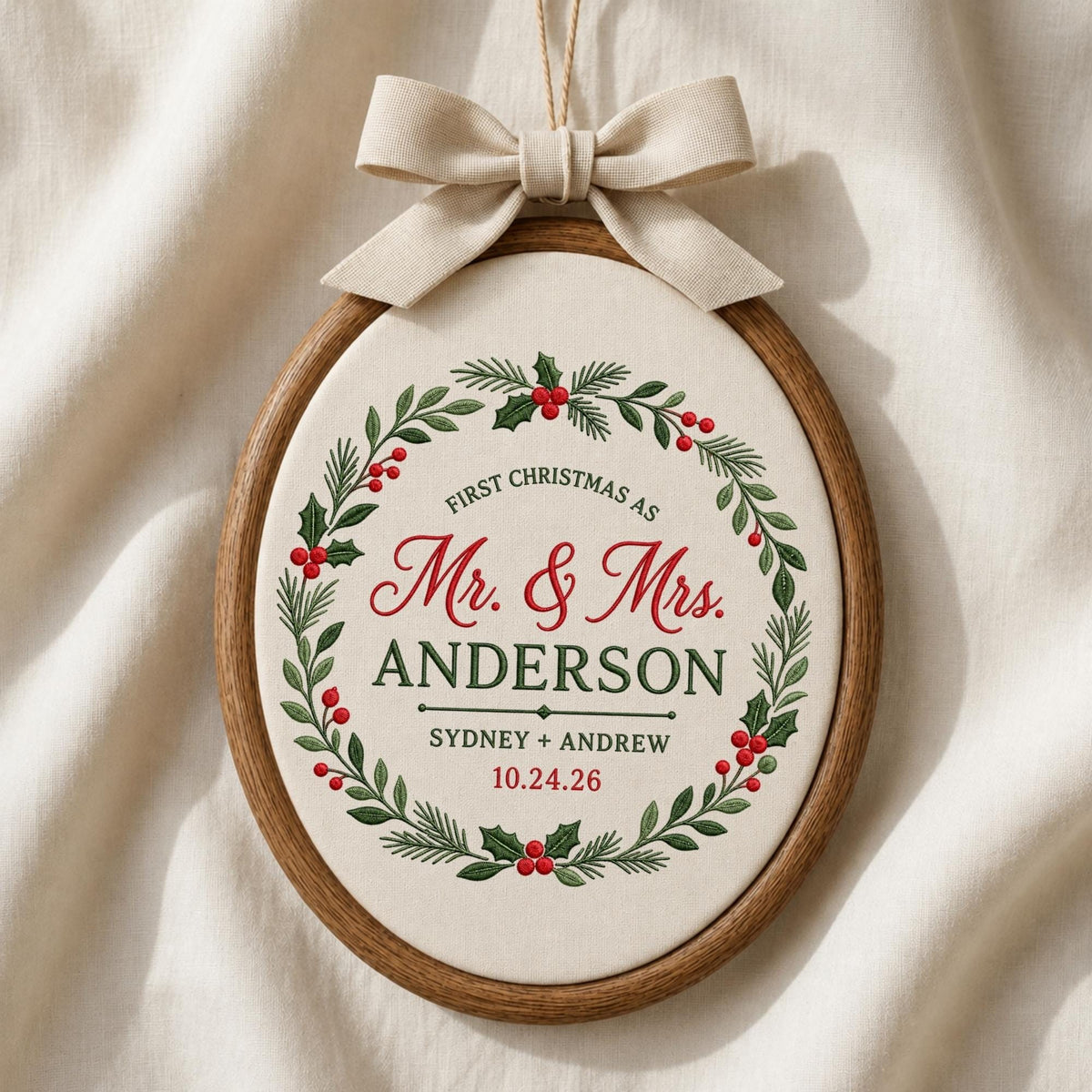 May include: An oval wooden embroidery hoop containing cream fabric with a green and red holly wreath design. The text reads: FIRST CHRISTMAS AS Mr. &amp; Mrs. ANDERSON SYDNEY + ANDREW 10.24.26. A beige fabric bow is attached to the top of the hoop.