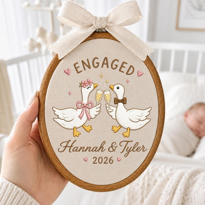 May include: An oval embroidery hoop featuring a beige fabric background with a cream-colored ribbon bow at the top. The design shows two white geese clinking champagne glasses, with the text "ENGAGED", "Hannah &amp; Tyler", and "2026" in brown.