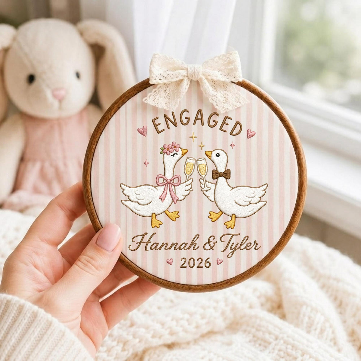 May include: A circular wooden embroidery hoop frame containing fabric with a light pink and white striped pattern. The design features two white geese clinking champagne glasses, the text ENGAGED, Hannah &amp; Tyler, and 2026, topped with a lace bow.