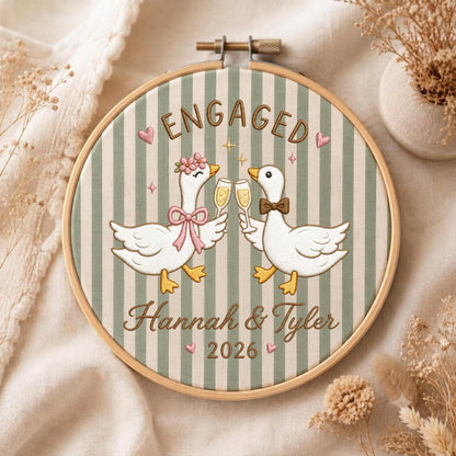 May include: A circular embroidery hoop featuring a green and white striped fabric background. Two white geese are embroidered in the center, one wearing a pink flower crown and bow, the other a brown bowtie. They hold champagne flutes. Text reads ENGAGED, Hannah &amp; Tyler, and 2026.