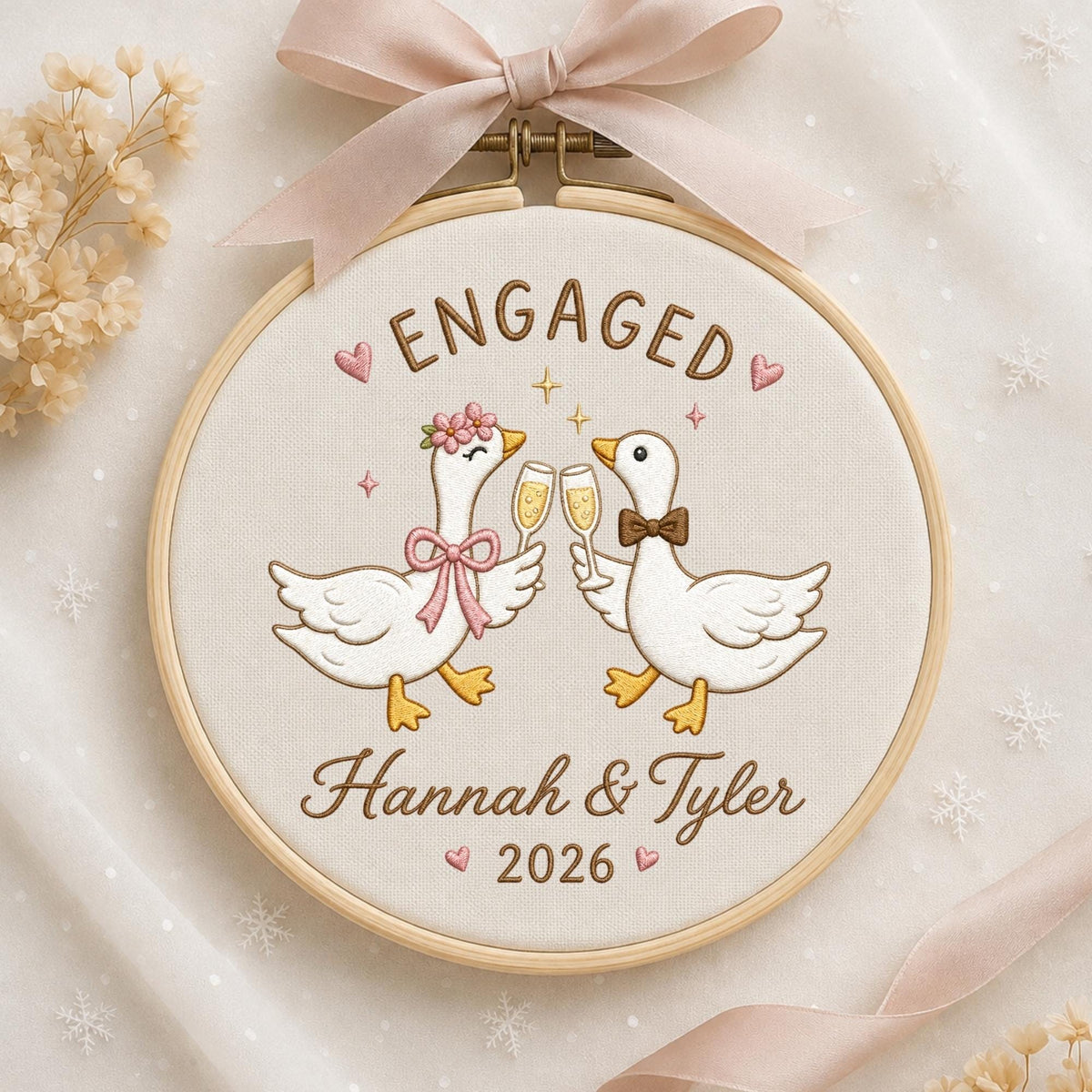 May include: A round wooden embroidery hoop containing white fabric with an embroidered design of two white geese clinking champagne glasses. The text reads ENGAGED, Hannah &amp; Tyler, and 2026. A light pink ribbon is tied at the top of the hoop.