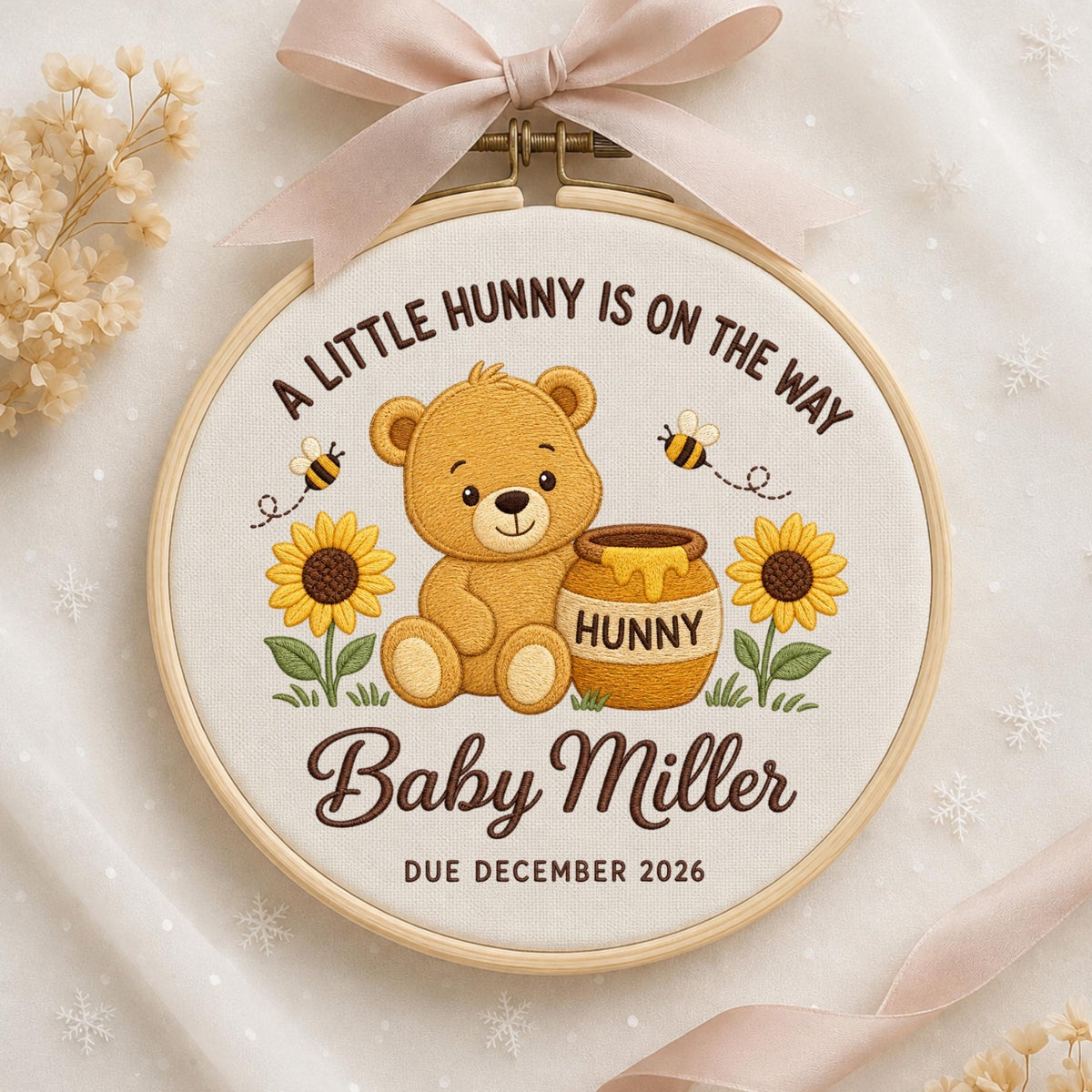 May include: A circular embroidery hoop featuring a light brown teddy bear sitting next to a honey pot labeled HUNNY, flanked by two sunflowers and bees. The text reads A LITTLE HUNNY IS ON THE WAY, Baby Miller, and DUE DECEMBER 2026, with a pink ribbon.