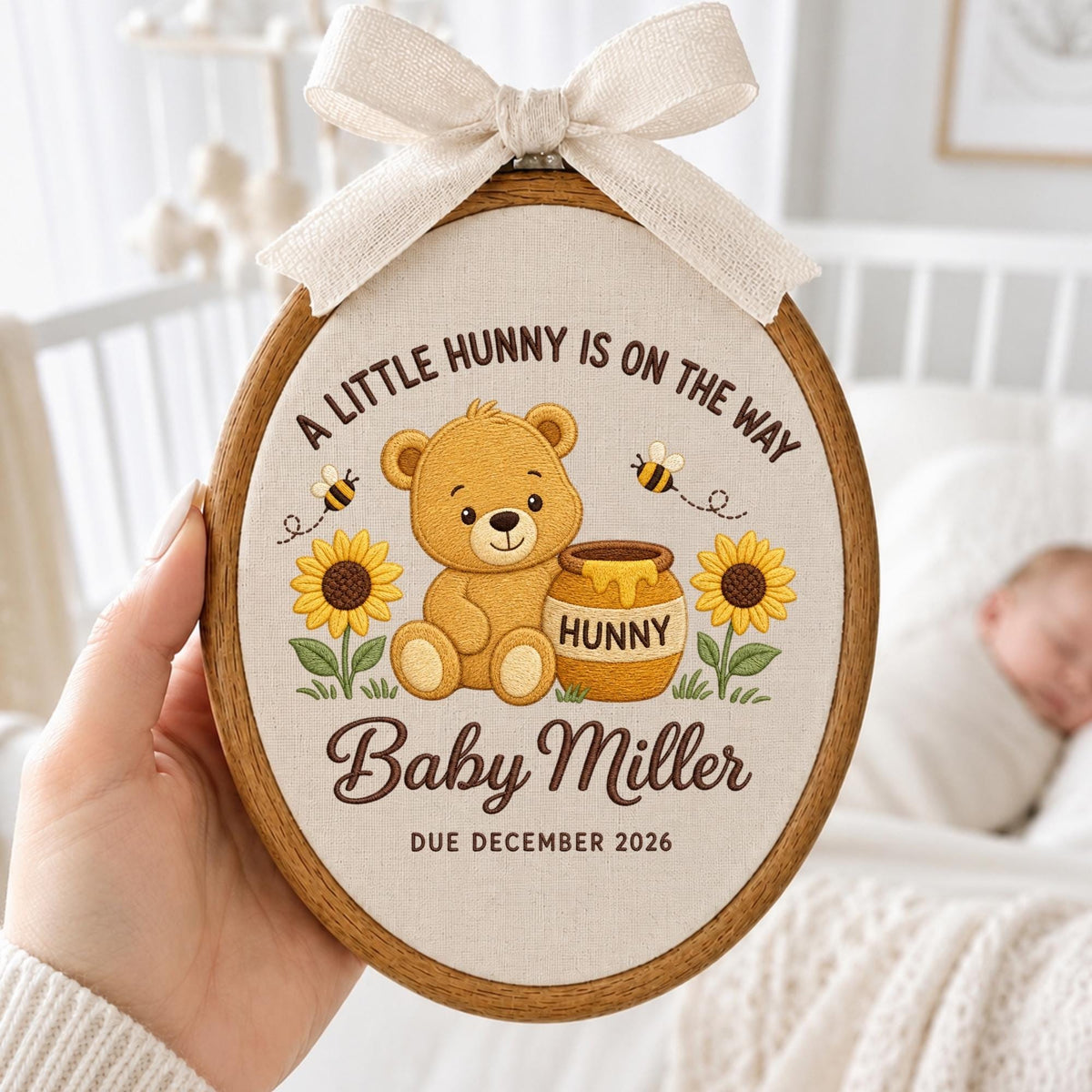 May include: An oval wooden embroidery hoop containing fabric with a brown bear, a honey pot labeled HUNNY, two sunflowers, and two bees. The text reads A LITTLE HUNNY IS ON THE WAY, Baby Miller, and DUE DECEMBER 2026. A cream ribbon bow is at the top.
