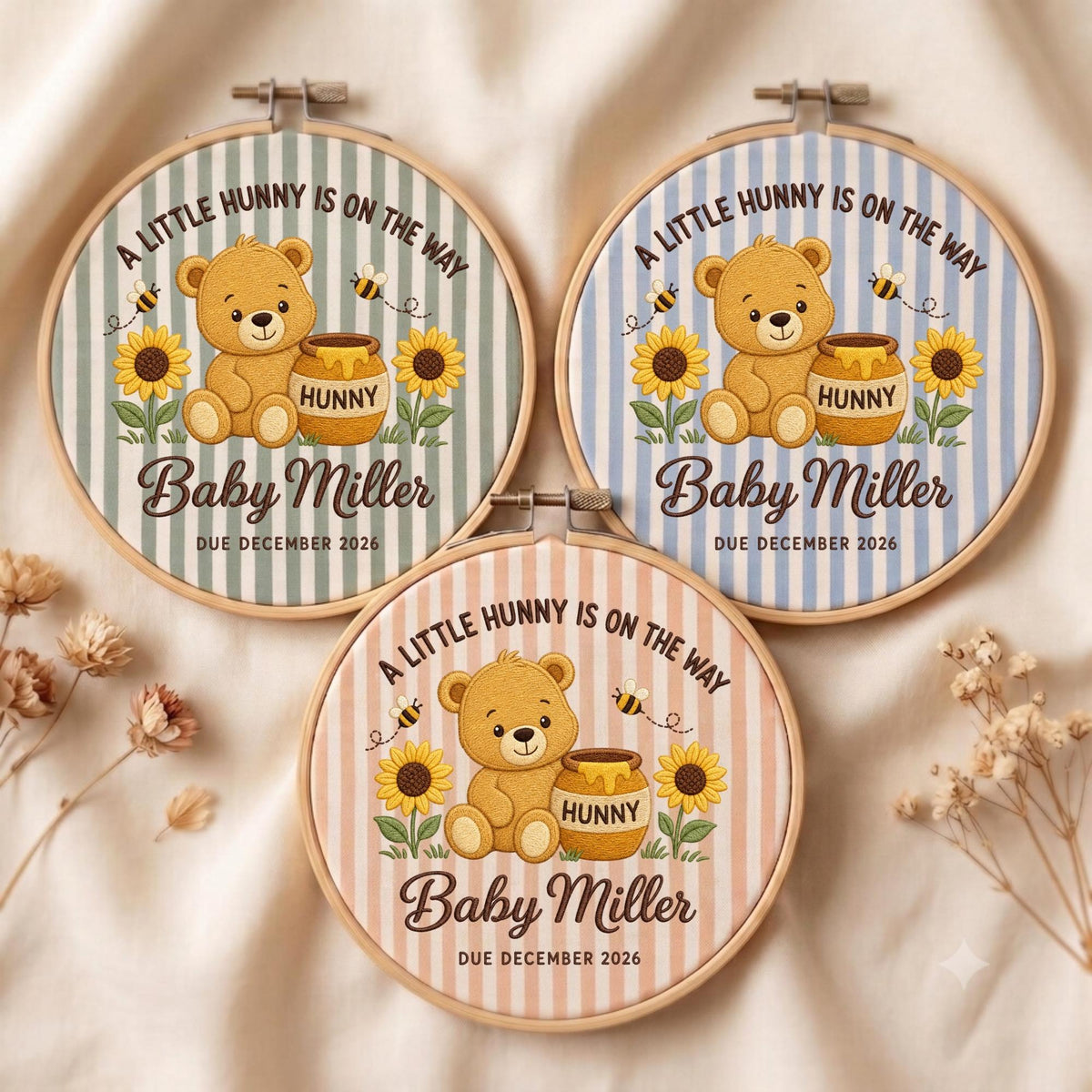 May include: Three circular wooden embroidery hoops featuring fabric with vertical stripes in sage green, light blue, and soft peach. Each displays a brown teddy bear, a honey pot labeled "HUNNY," sunflowers, bees, and the text "A LITTLE HUNNY IS ON THE WAY Baby Miller DUE DECEMBER 2026."