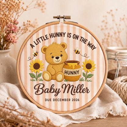 May include: A round wooden embroidery hoop containing fabric with vertical light pink stripes. The design features a brown teddy bear, a honey pot labeled HUNNY, two sunflowers, and bees. Text reads: A LITTLE HUNNY IS ON THE WAY, Baby Miller, DUE DECEMBER 2026.