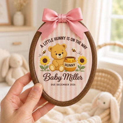 May include: An oval wooden embroidery hoop frame containing fabric with a pink and white striped pattern. The design features a brown teddy bear, a honey pot labeled "HUNNY," sunflowers, bees, and the text "A LITTLE HUNNY IS ON THE WAY," "Baby Miller," and "DUE DECEMBER 2026." A pink satin bow is attached to the top.