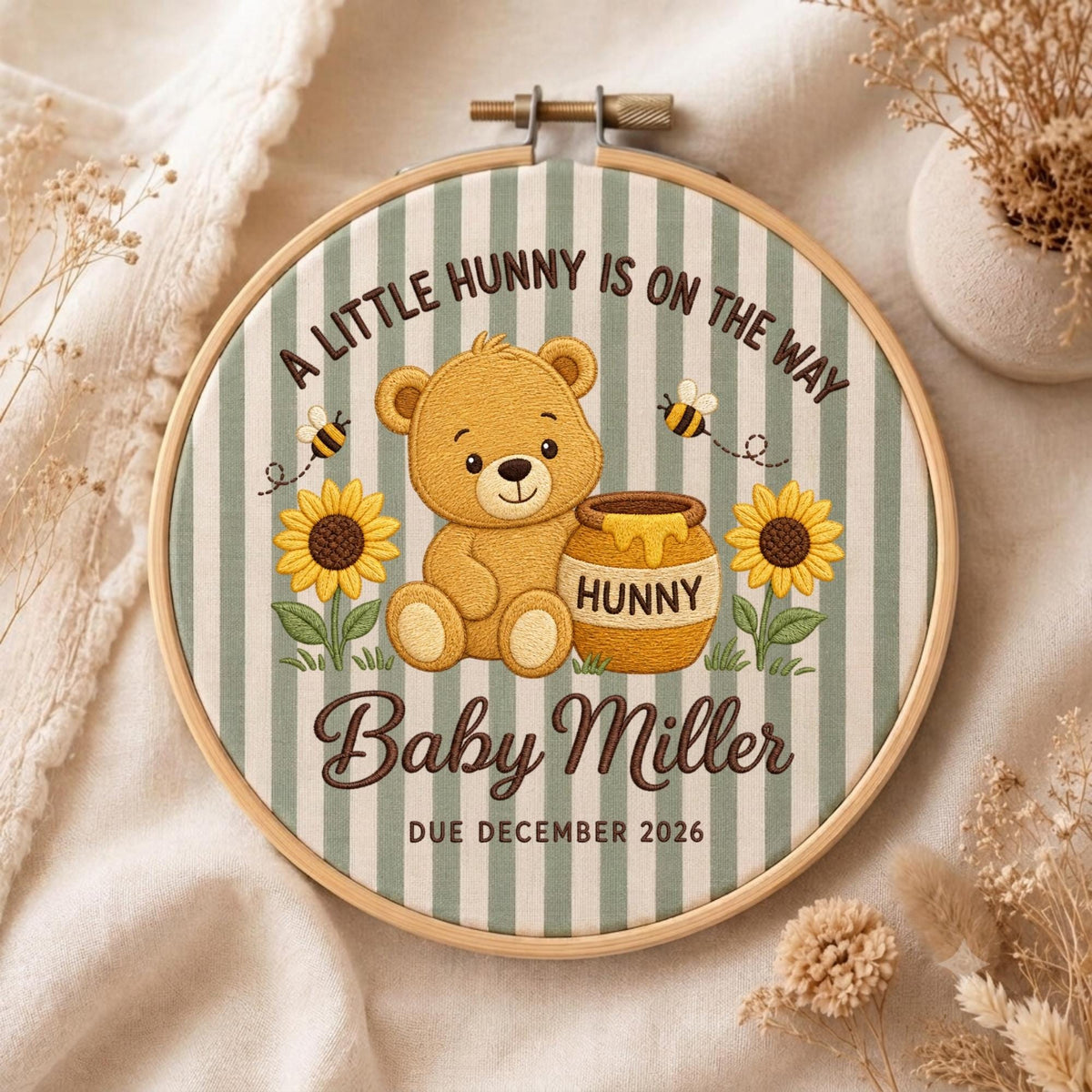 May include: A round wooden embroidery hoop containing fabric with green and white vertical stripes. The embroidery features a brown teddy bear, a honey pot labeled HUNNY, two sunflowers, and two bees. Text reads: A LITTLE HUNNY IS ON THE WAY, Baby Miller, DUE DECEMBER 2026.