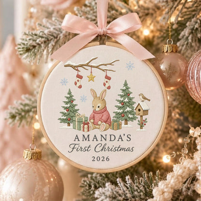 May include: A round wooden embroidery hoop ornament featuring a light-colored fabric with a printed illustration of a rabbit in a pink sweater, Christmas trees, gifts, and stockings. The text reads AMANDA'S First Christmas 2026. A pink ribbon bow is attached.