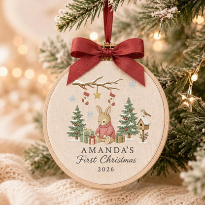 May include: A round wooden embroidery hoop ornament featuring a beige fabric center with a printed illustration of a rabbit in a pink sweater, Christmas trees, gifts, and stockings. A dark red ribbon bow is tied at the top with the text AMANDA'S First Christmas 2026.