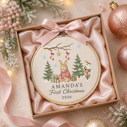 May include: A round wooden embroidery hoop containing a fabric panel with a printed illustration of a rabbit in a pink sweater, Christmas trees, gifts, and stockings. The text reads AMANDA'S First Christmas 2026. A pink satin bow is attached.