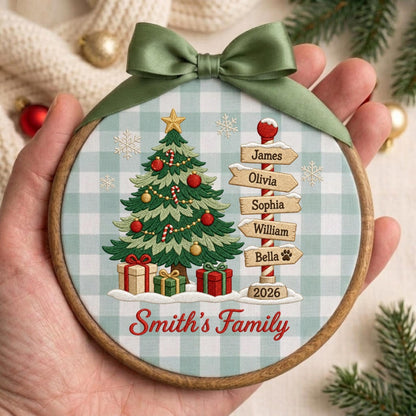 May include: A round wooden embroidery hoop ornament featuring a light blue and white gingham fabric background. It displays an embroidered Christmas tree with gifts, a signpost with names James, Olivia, Sophia, William, Bella, and the year 2026.