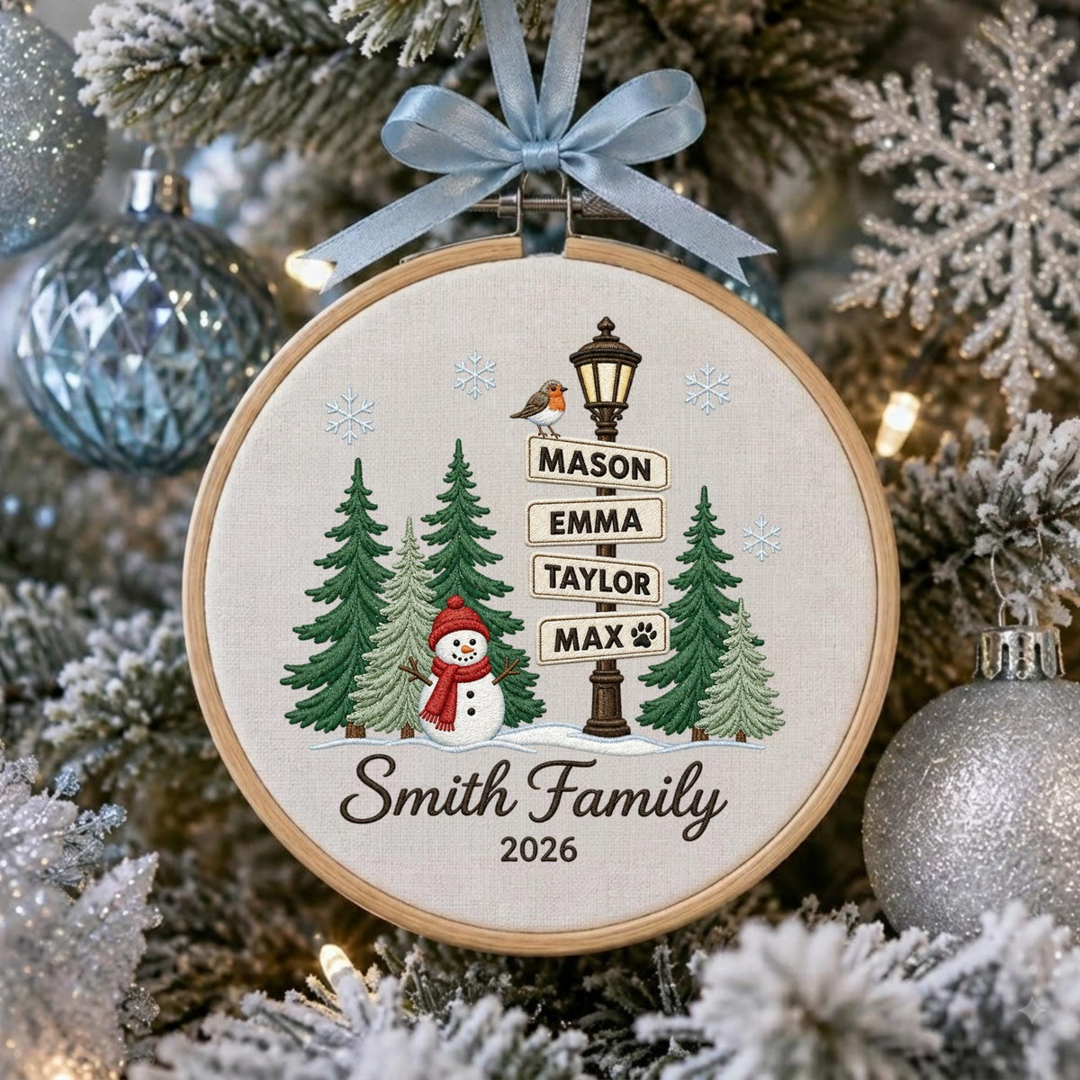 May include: A round wooden embroidery hoop ornament featuring a light-colored fabric background. The design includes a snowman, green pine trees, a street lamp with signs reading Mason, Emma, Taylor, Max, and the text Smith Family 2026 with a blue ribbon.