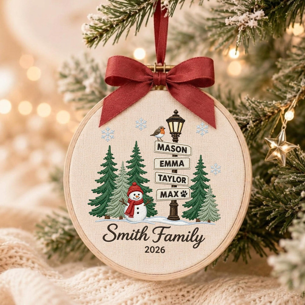 May include: A round wooden embroidery hoop ornament featuring a cream fabric background with a printed winter scene of a snowman, green pine trees, and a street sign listing the names Mason, Emma, Taylor, and Max. It has a dark red ribbon bow.
