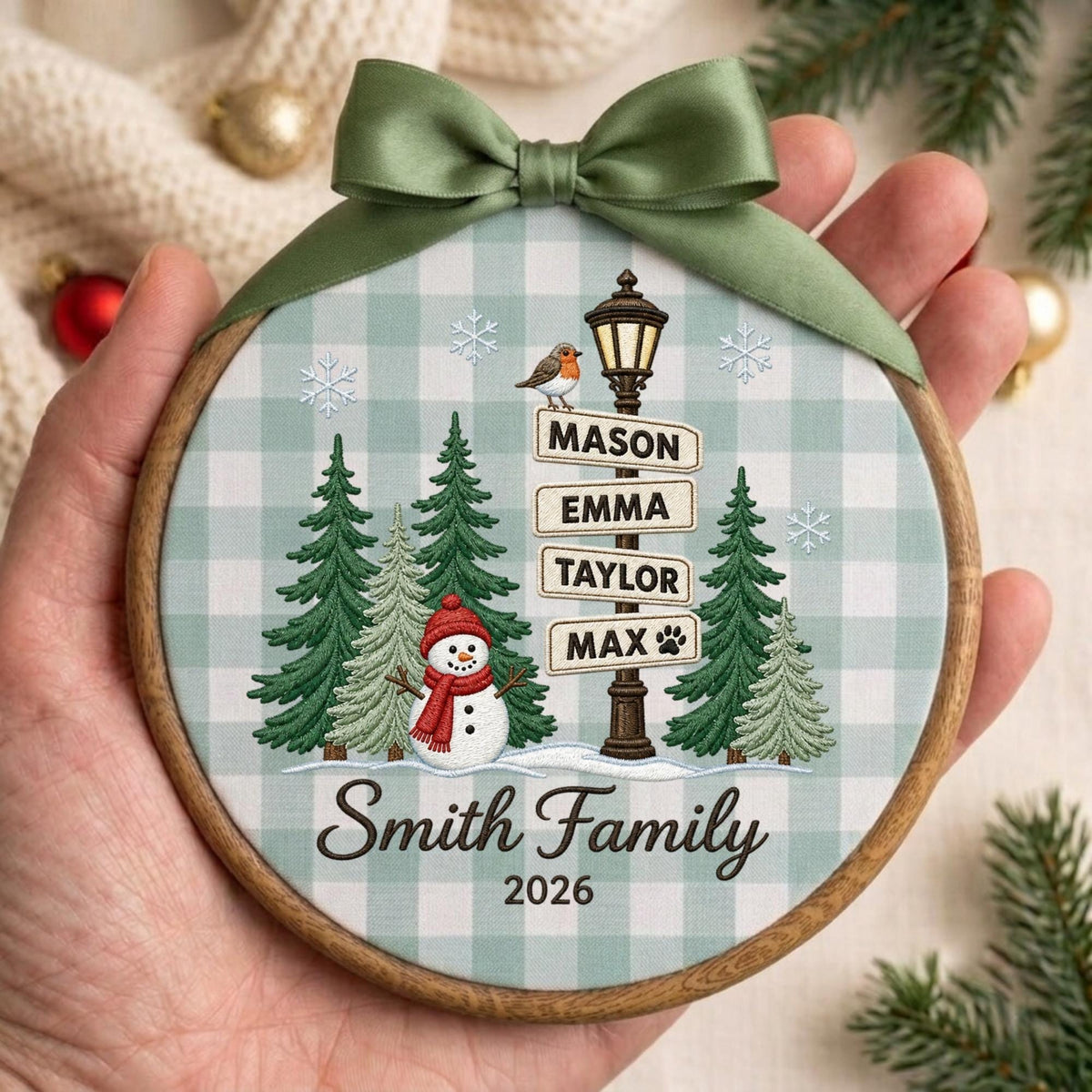 May include: A round wooden embroidery hoop ornament featuring a light green and white buffalo check background. It displays a snowman, pine trees, a street sign with the names Mason, Emma, Taylor, Max, and the text Smith Family 2026 with a green bow.