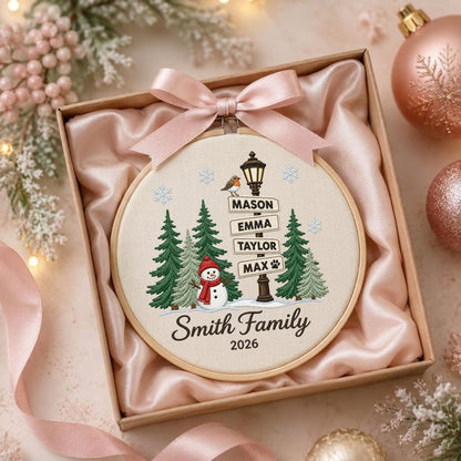 May include: A circular embroidery hoop featuring a winter scene with a snowman, evergreen trees, and a street lamp with signs reading MASON, EMMA, TAYLOR, and MAX. The text Smith Family 2026 is embroidered below. A light pink satin ribbon is tied at the top.