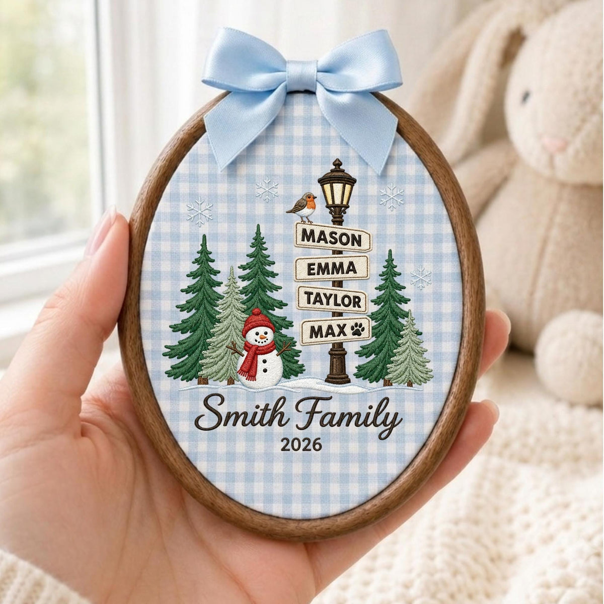 May include: An oval wooden embroidery hoop ornament featuring a light blue gingham fabric background. The design includes a snowman, green pine trees, a street lamp with signs reading Mason, Emma, Taylor, Max, and the text Smith Family 2026.