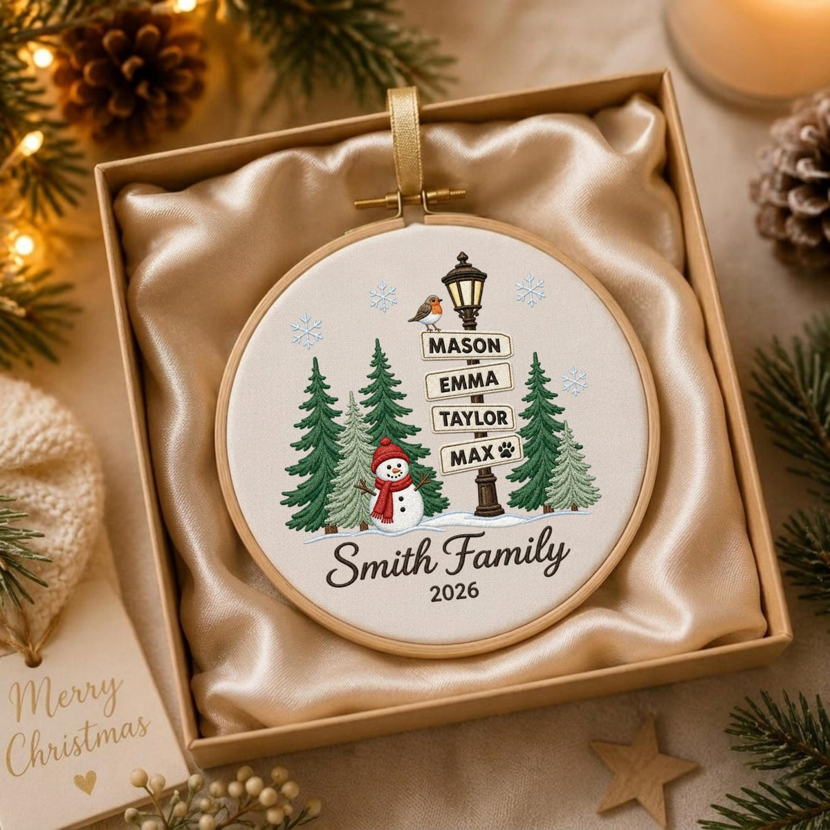 May include: A round wooden embroidery hoop ornament featuring a fabric insert with a printed winter scene. The design includes a black lamppost with signs reading Mason, Emma, Taylor, and Max, a snowman, pine trees, snowflakes, and the text Smith Family 2026.