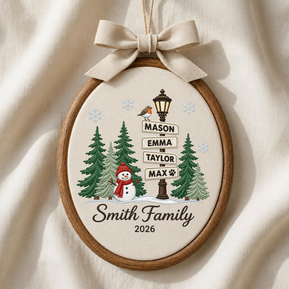 May include: An oval embroidery hoop ornament featuring a winter scene with a snowman, pine trees, and a street sign listing the names Mason, Emma, Taylor, and Max with a paw print. The text Smith Family 2026 is below, topped with a beige fabric bow.
