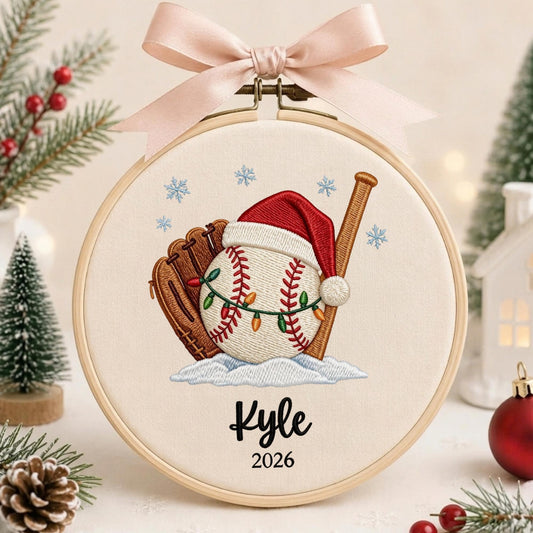 May include: A round wooden embroidery hoop containing cream-colored fabric with an embroidered baseball, brown glove, and wooden bat. The baseball wears a red Santa hat and string lights. Below, the name Kyle and year 2026 are stitched in black.