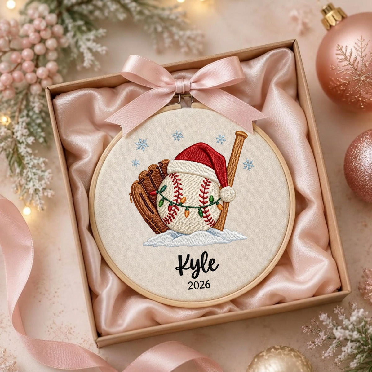 May include: A round wooden embroidery hoop containing a fabric panel with an embroidered baseball, glove, and bat. The baseball wears a red Santa hat and is wrapped in string lights. The text Kyle and 2026 is embroidered below. A pink satin bow is attached.