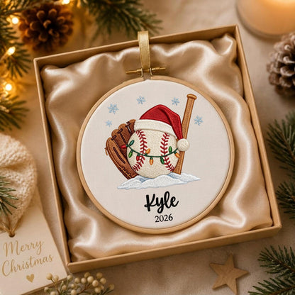 May include: A round embroidery hoop ornament featuring a baseball wearing a red Santa hat, a brown baseball glove, and a wooden bat. The baseball is wrapped in colorful string lights, set against a white background with blue snowflakes, the name Kyle, and the year 2026.