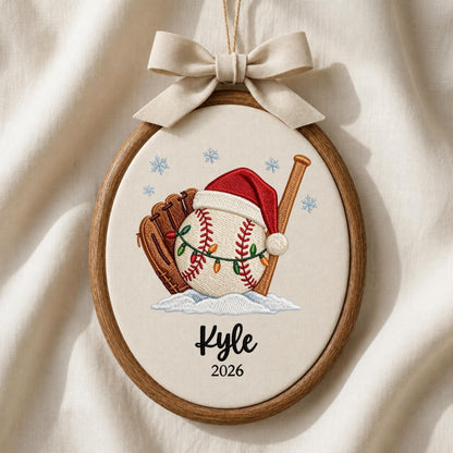May include: An oval wooden embroidery hoop ornament featuring a baseball with a Santa hat and string lights, a brown glove, and a wooden bat. The design includes blue snowflakes, the name Kyle, and the year 2026, topped with a beige fabric bow.