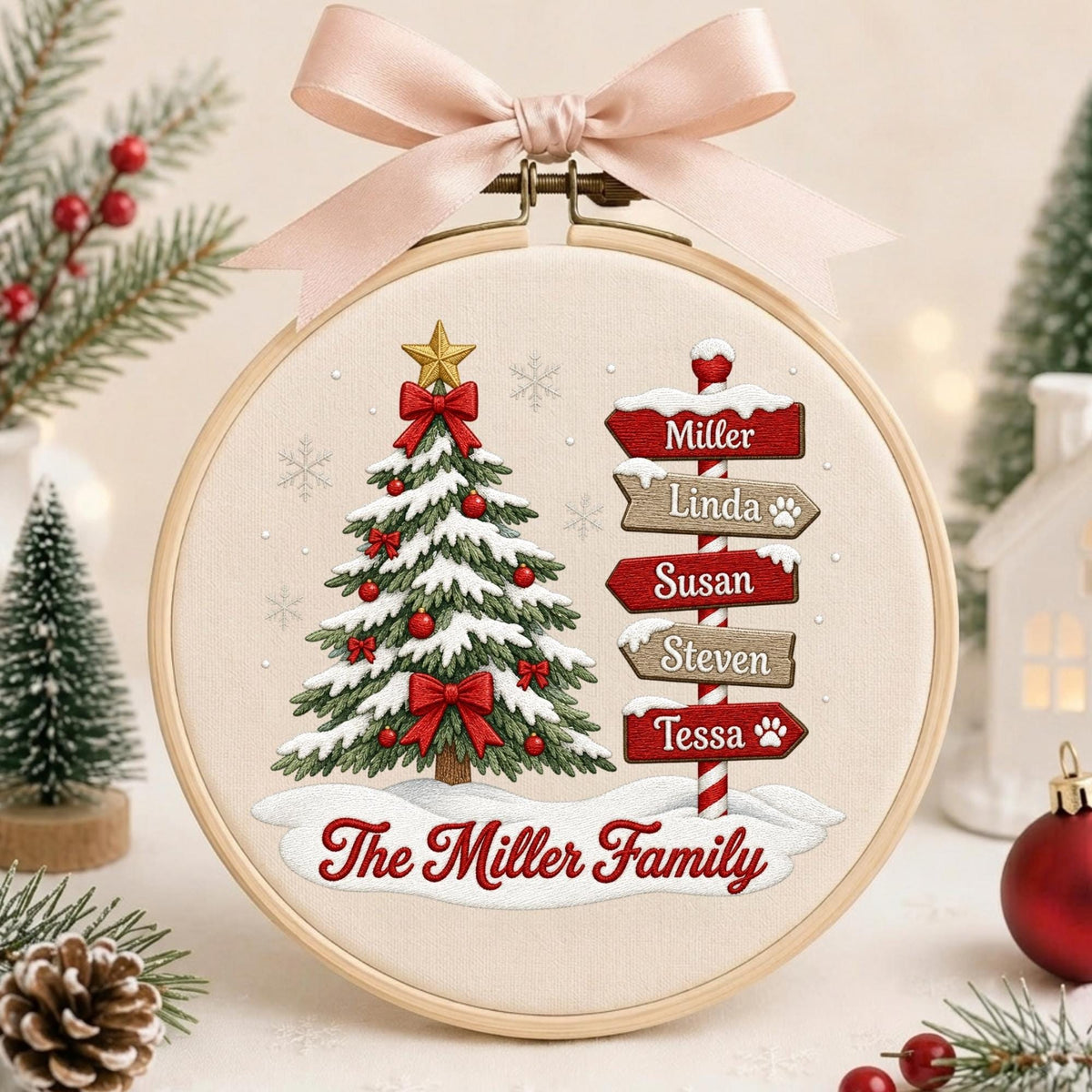 May include: A circular embroidery hoop featuring a Christmas tree with snow, red bows, and a gold star. To the right, a signpost displays the names Miller, Linda, Susan, Steven, and Tessa. The text The Miller Family is printed below, topped with a pink bow.