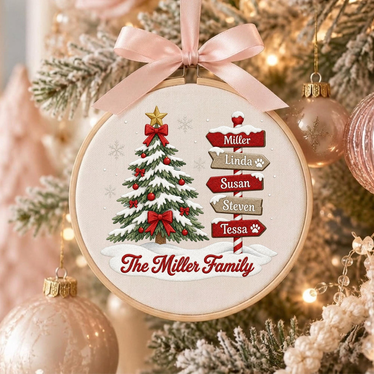 May include: A round embroidery hoop ornament featuring a Christmas tree with red bows and a gold star, next to a signpost with the names Miller, Linda, Susan, Steven, and Tessa. The text The Miller Family is printed below in red script.