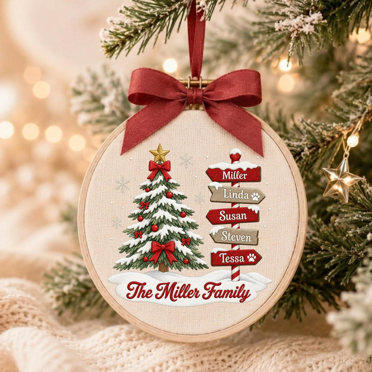 May include: A round embroidery hoop ornament featuring a Christmas tree with red ornaments and a gold star, next to a signpost with the names Miller, Linda, Susan, Steven, and Tessa. The text The Miller Family is printed below in red.