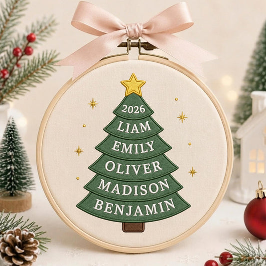 May include: A circular wooden embroidery hoop containing a cream-colored fabric with a green embroidered Christmas tree. The tree features a yellow star at the top and the text 2026, Liam, Emily, Oliver, Madison, and Benjamin. A pink ribbon is tied at the top.
