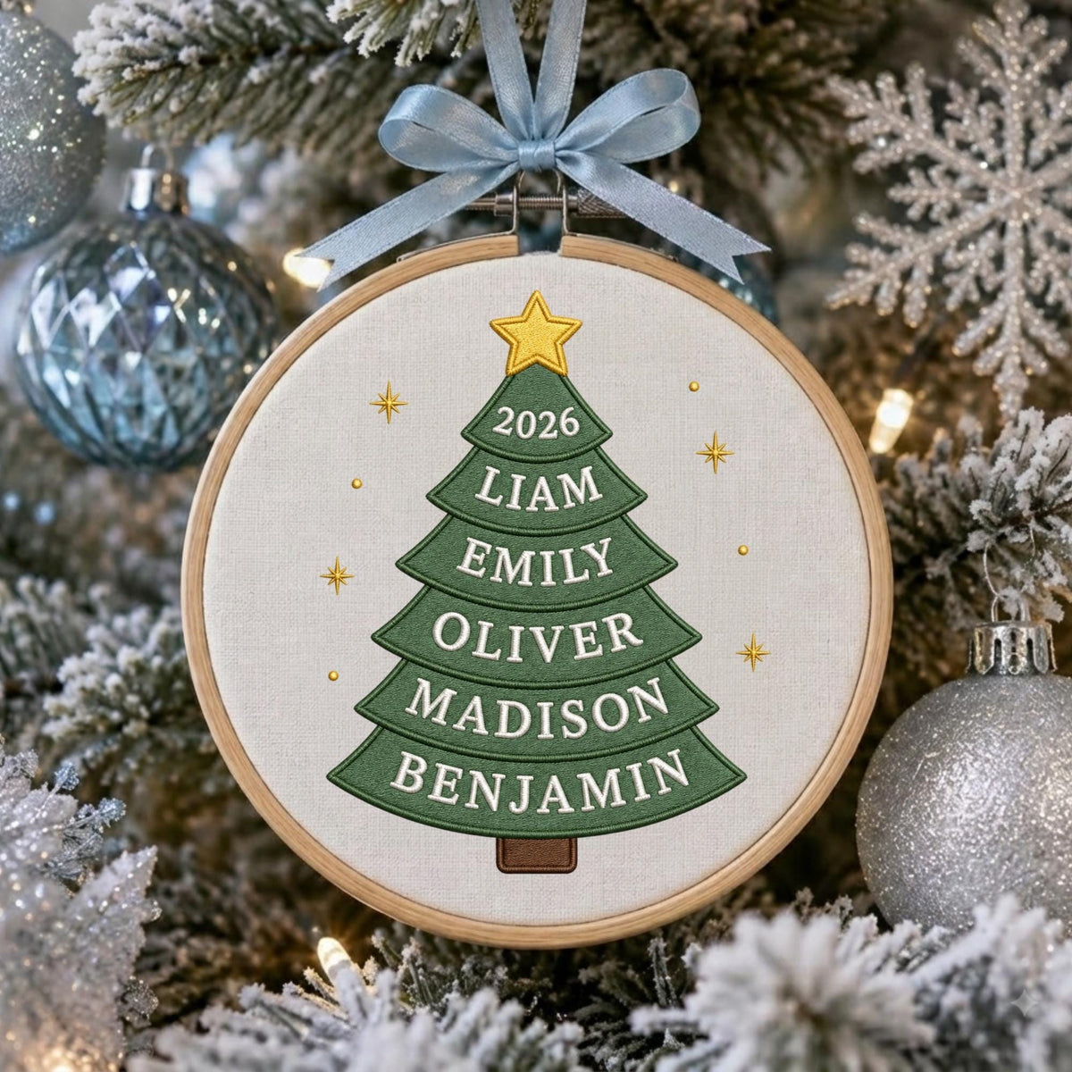May include: A circular wooden embroidery hoop frame containing a fabric panel with an embroidered green Christmas tree. The tree features a yellow star at the top and the text 2026, LIAM, EMILY, OLIVER, MADISON, and BENJAMIN. A light blue ribbon bow is attached to the top.