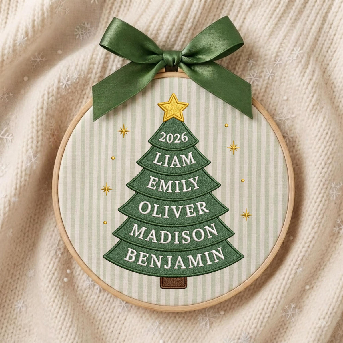 May include: A circular wooden embroidery hoop containing fabric with a vertical striped pattern. A green embroidered Christmas tree features a yellow star at the top and the text 2026, LIAM, EMILY, OLIVER, MADISON, BENJAMIN. A dark green satin ribbon bow is attached to the top.