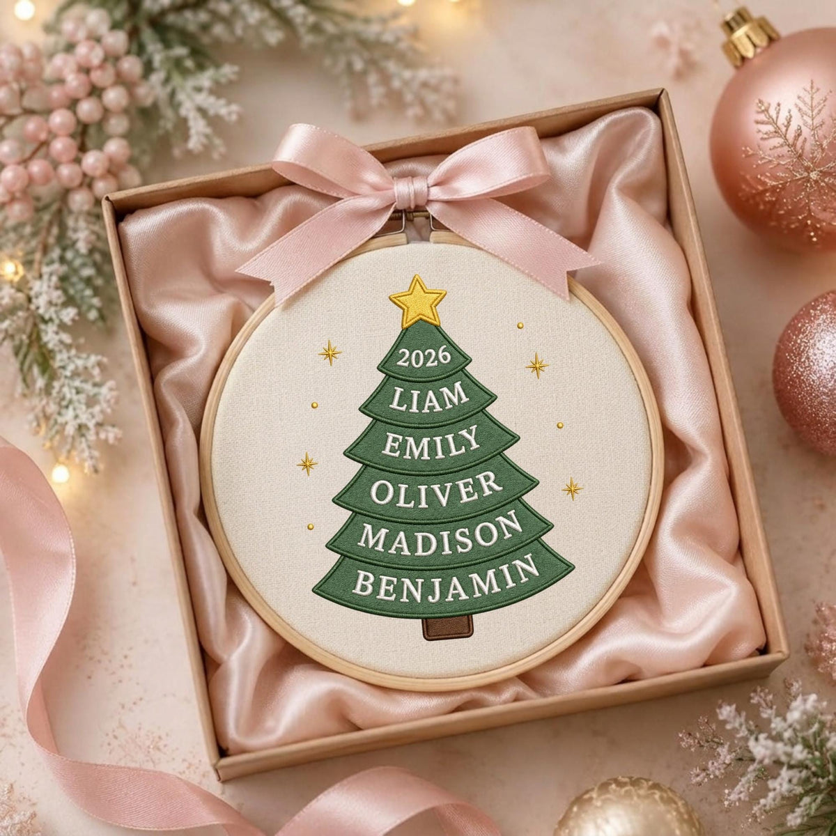 May include: A circular embroidery hoop featuring a green fabric Christmas tree with a yellow star on top. The tree is embroidered with the year 2026 and the names Liam, Emily, Oliver, Madison, and Benjamin. A light pink satin ribbon bow is attached.