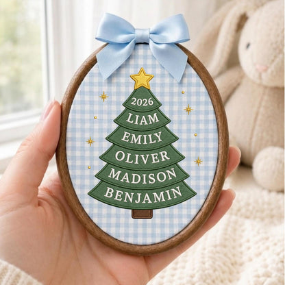 May include: An oval wooden embroidery hoop ornament featuring a light blue and white gingham fabric background. A green Christmas tree graphic is centered with a yellow star on top and the text 2026, LIAM, EMILY, OLIVER, MADISON, BENJAMIN. A light blue satin bow is attached to the top.