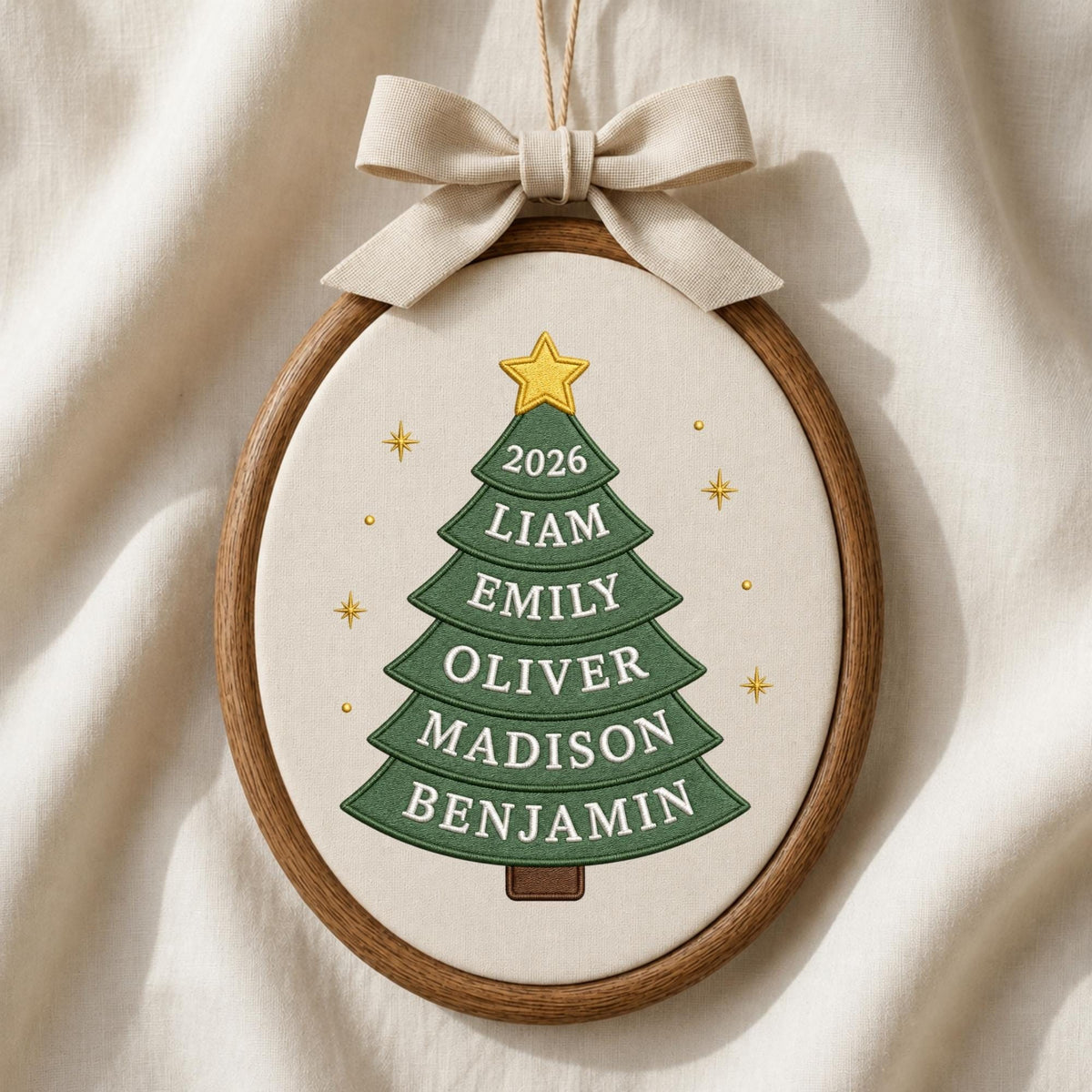 May include: An oval embroidery hoop ornament featuring a green fabric Christmas tree with a yellow star on top. The tree displays the year 2026 and the names Liam, Emily, Oliver, Madison, and Benjamin. A beige fabric bow is attached to the top.