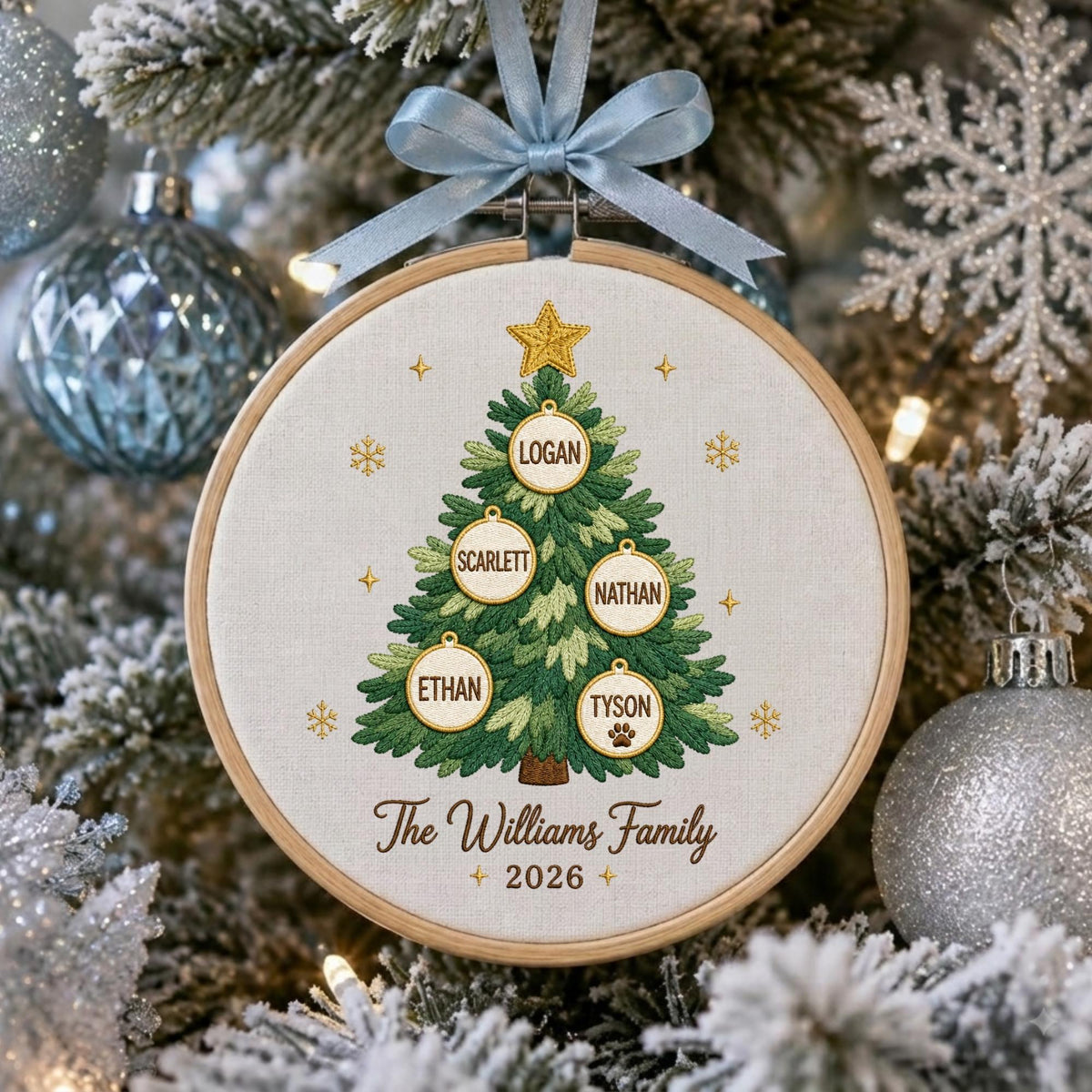May include: A round wooden embroidery hoop ornament featuring a stitched green Christmas tree with a gold star. Five white circular ornaments hang on the tree with the names Logan, Scarlett, Nathan, Ethan, and Tyson. Text reads The Williams Family 2026.