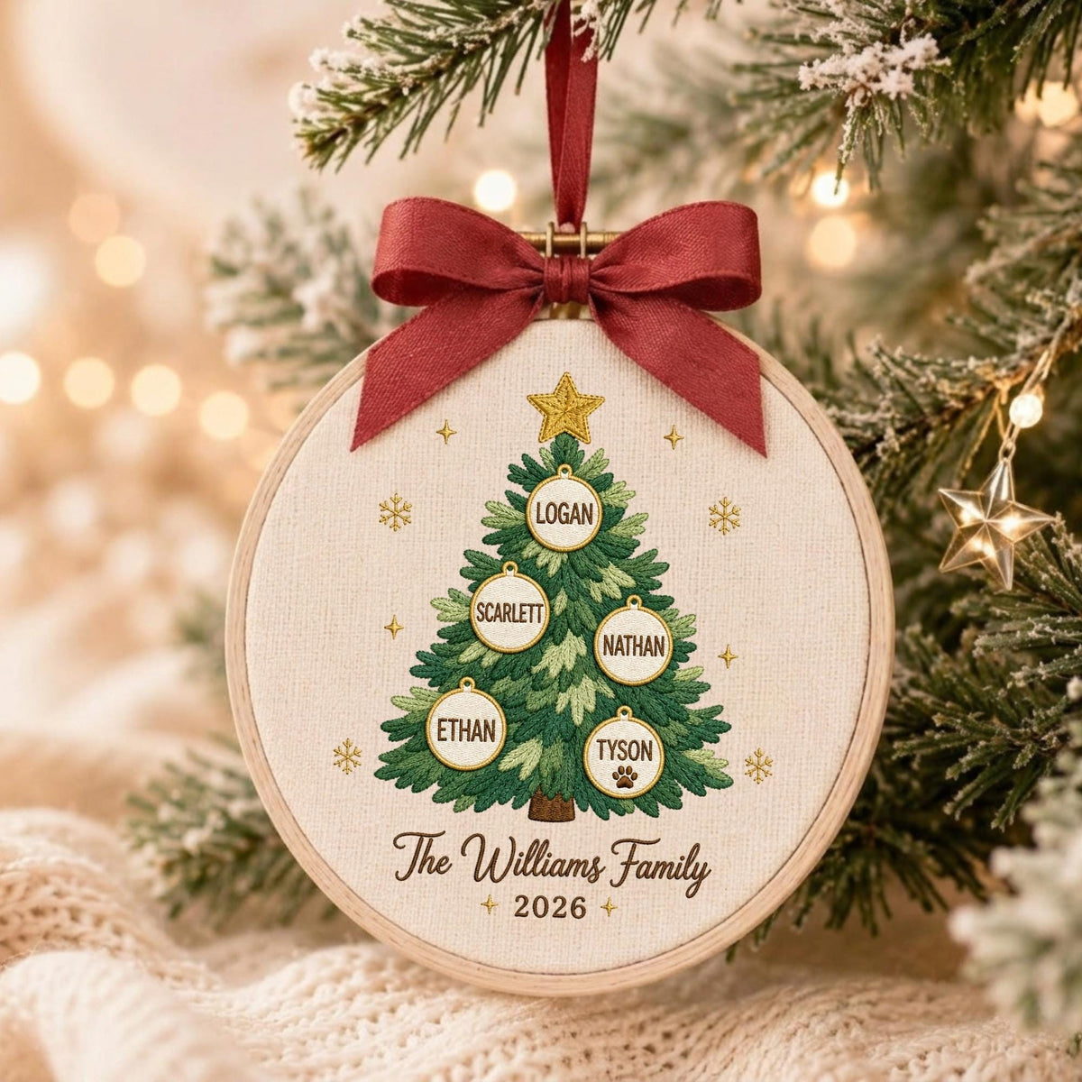May include: A round wooden embroidery hoop ornament featuring a stitched green Christmas tree with a gold star on top. Five white circular ornaments hang on the tree with the names Logan, Scarlett, Nathan, Ethan, and Tyson. Text reads The Williams Family 2026.